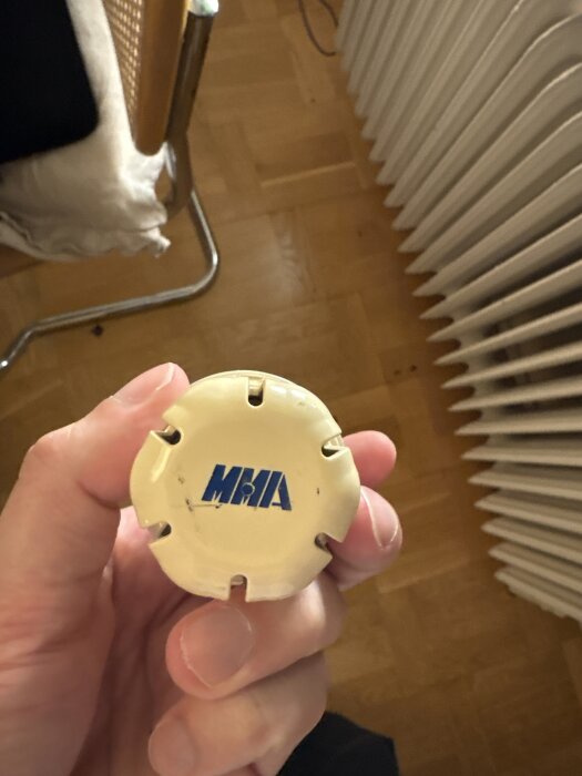 Hand som håller en vit termostat med texten "MMA", en stol och ett element syns i bakgrunden.