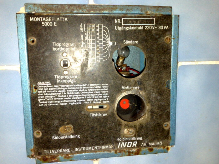 Kontrollpanel för automatisk spolning av pissoarer, med justeringar och märkningar, monterad på en blå kakelvägg.