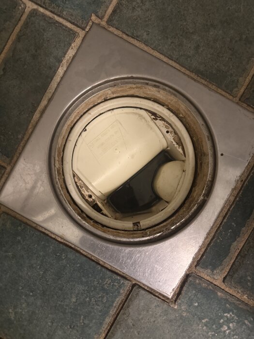 Duschgolvbrunn med synlig klämring som sitter löst, omgiven av blå-gröna kakelplattor, potentiellt från byggåret 1991.