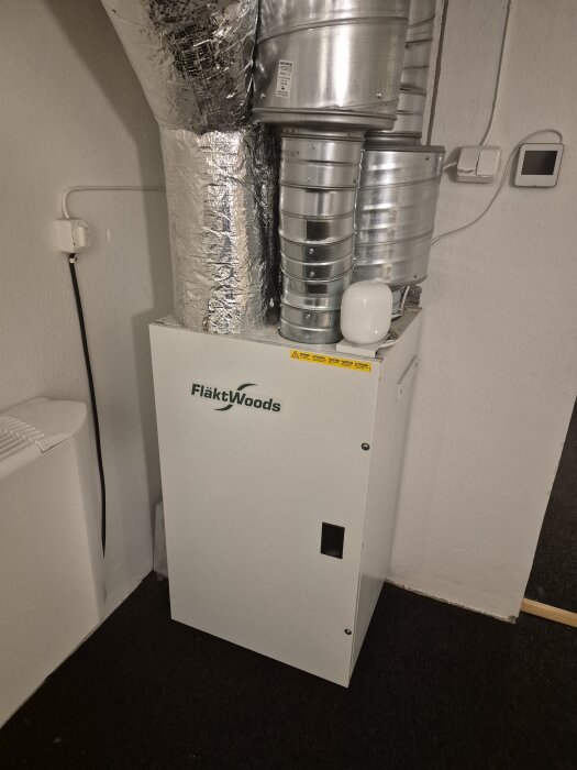 Vit ventilationsenhet med rör och isolering, märket Fläkt Woods, i ett hushållsutrymme.