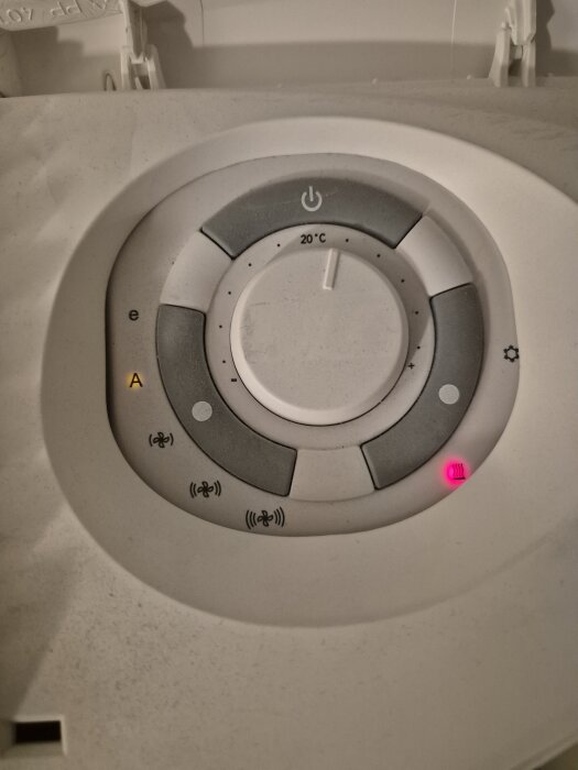 Rund termostatpanel med temperaturinställning och olika symboler för värme, fläkt och luftkvalitet. Inställt på 20°C med en rosa lysande indikation.
