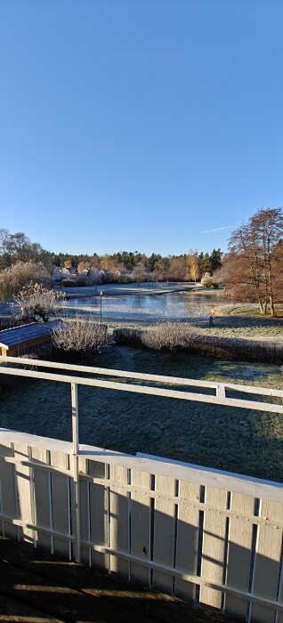 Utsikt från sovrummet över en frusen sjö med omgivande gräsmattor och träd, under en klarblå himmel.