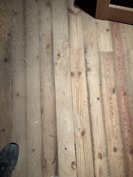 Ojämnt och slitet träplankgolv med kvistar och springor i ett äldre hus, fotat ovanifrån, delvis skymd av en sko.