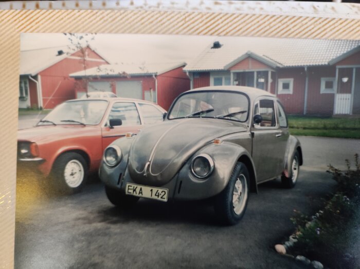 Grå klassisk bil parkerad framför en röd bil på en gata med röda småhus i bakgrunden, registreringsnummer EKA 142.