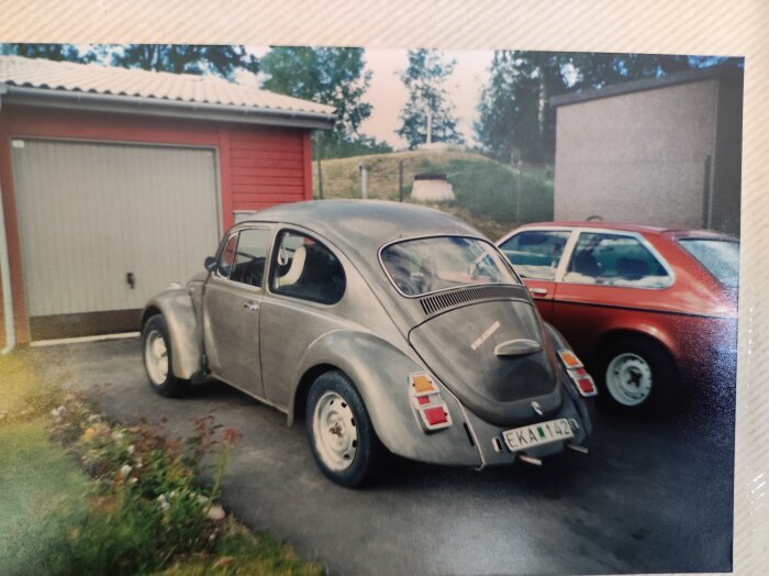 Svart veteranbil parkerad bredvid en röd bil framför ett garage. Tre olika bakljus har monterats på den bakre stötfångaren.