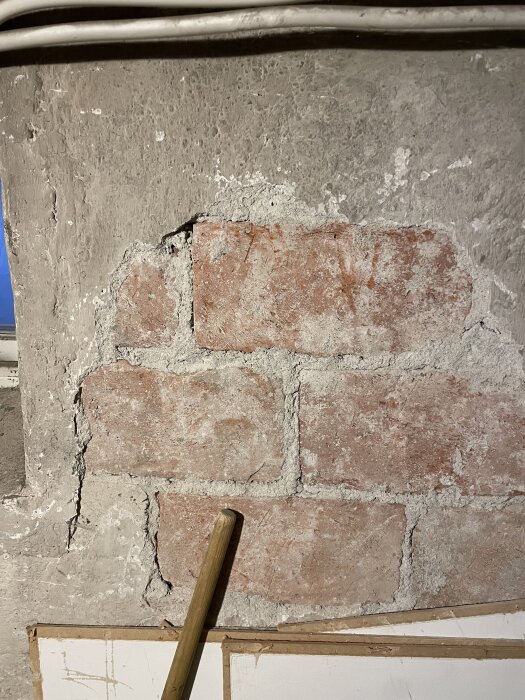 Skadad innervägg i källare med synliga tegelstenar, gammal puts och hålrum, samt en borr stående mot väggen.