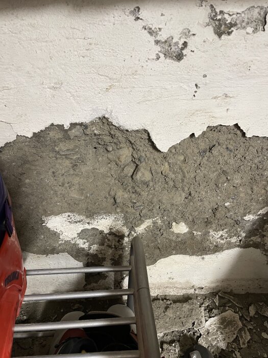 Skadad källarvägg med vit puts som flagnar, exponerar ojämn betong med stora stenar. Röd föremål och metallställning syns nedtill.