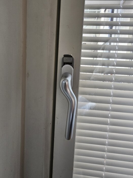 Balkongdörrens handtag i låst läge, delvis täckt av persienner.