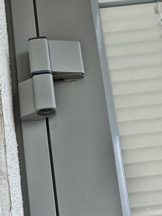 Bilden visar ett gångjärn på en balkongdörr i närbild, med grå metallfinish och en del av dörrkarmen synlig.