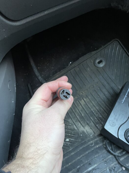 Hand som håller en kontakt med två stift i en bils fotutrymme, ovanpå en gummimatta.