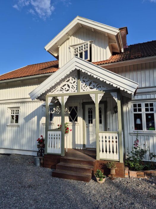 Vit farstukvist med snickarglädje och röda pelargoner framför trähus, blå himmel i bakgrunden.