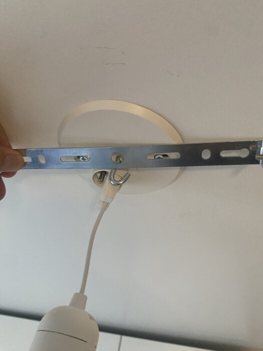 Metallhållare monterad ovanpå en vit takinfattning med en krok och elektrisk ledning synlig.