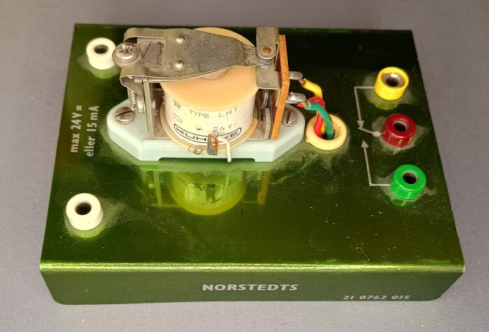 Elektriskt relä från undervisningsmaterial på sjuttiotalet, placerat på en grön plattform med märkningen 24V och olika färgade kontakter.