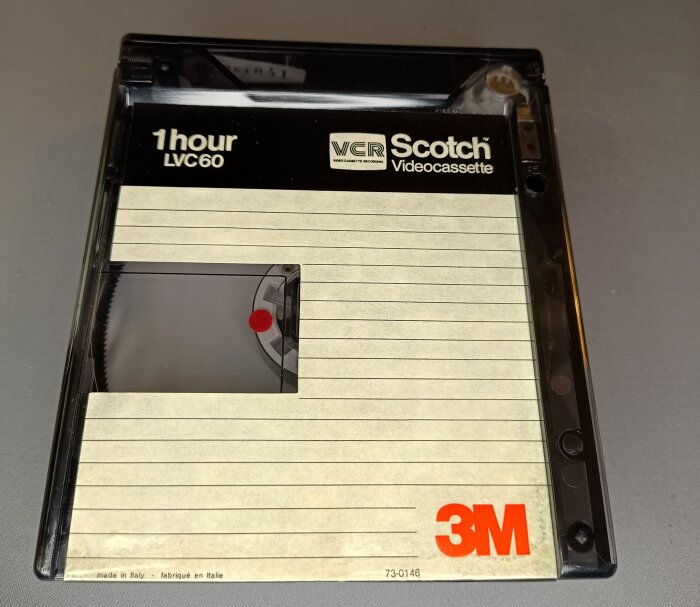 VCR-kassett från Scotch med 3M-logo och text “1 hour LVC60”, synligt kassettband genom plastfönster, placerad på en grå bakgrund.