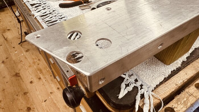 Aluminiumprofil i L-form med borrade hål, placerad på en arbetsbänk med trä- och tygdetaljer i bakgrunden.