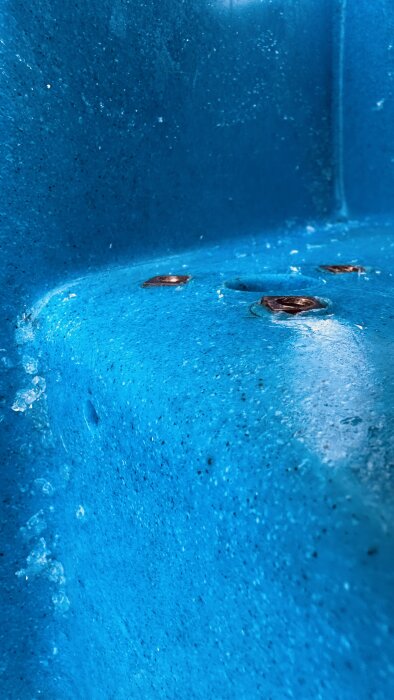 Närbild av blå plastyta med isbildning och metallfästen, från en trampbåt under vinterförvaring.