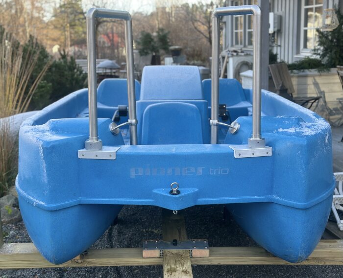 Blå trampbåt med två rostfria räcken, ståendes på trästöd utomhus, vinterförberedd.