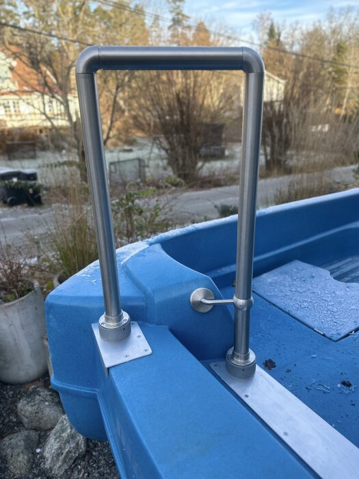 Rostfria räcken monterade på blå trampbåt, fästa vid aluminiumplattor. Bakgrund: träd och hus.