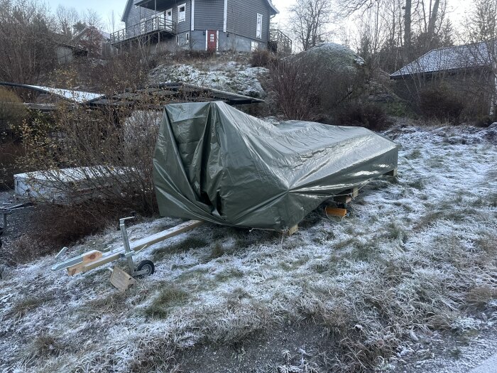 Trampbåt täckt med grön presenning på frostig mark, bredvid en båtvagn. Hus och vinterlandskap i bakgrunden.