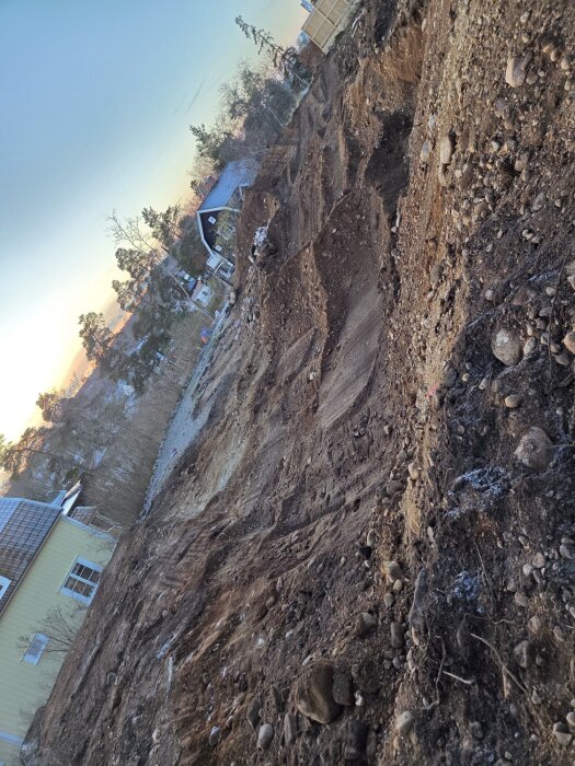 Grusig och stenig markmassa på en lutande tomt, med träd och hus i bakgrunden under en blåorange himmel.