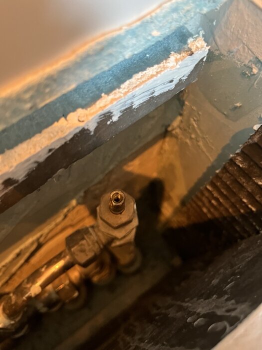 Gammal golvtermostat i metall inuti en vägg, troligen från ett hus byggt 1989, med kopparrör synliga.