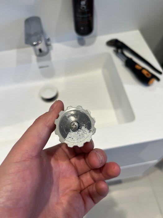 Hand håller en äldre golvtermostat med en tärningsliknande vridknapp framför en vit tvättställ. Verktyg syns i bakgrunden.