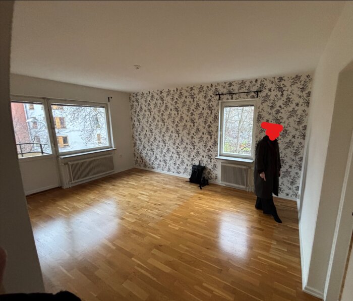 Ett rum med trägolv som ser slitet ut, tapetserade väggar och stora fönster. En person står vid en vägg.