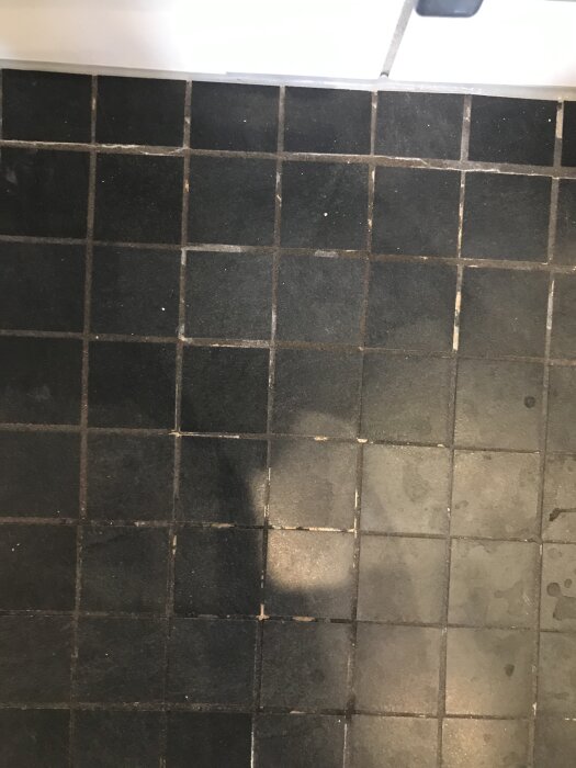Svarta golvklinkers med slitna och missfärgade fogar i ett badrum, speciellt synligt i duschhörnan.