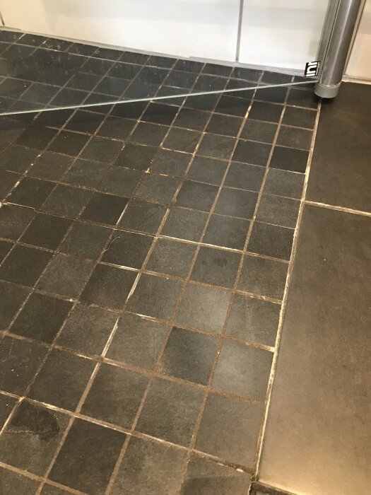 Svart badrumsgolv med kvadratiska klinkers; fogarna är slitna och missfärgade, särskilt i hörnet där duschens glasvägg syns.