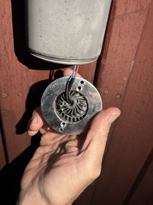 Hand håller en rund, metallisk ljuskälla med kablar som går upp mot ett grått hölje monterat på en röd trävägg.