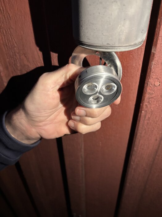 Hand håller en rund, metallisk utomhusbelysning med tre ljuskällor, monterad på en vägg.