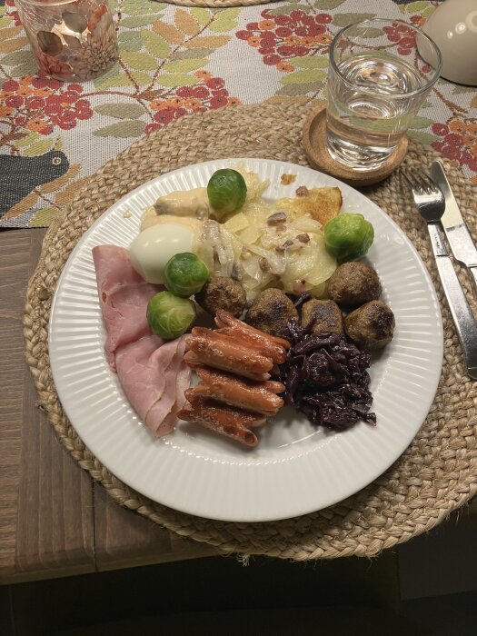 Tallrik med blandade rätter, inkl. brunkål, köttbullar, skinka, potatisgratäng, brysselkål och prinskorv, med ett glas vatten på ett dukat bord.