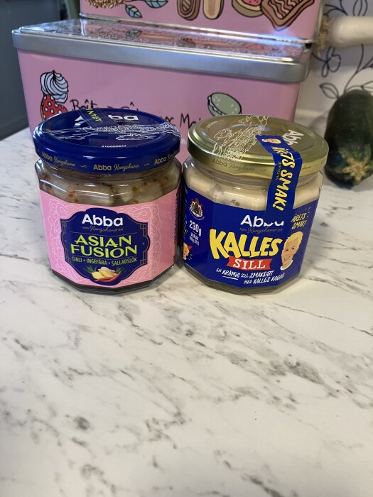 Två burkar sill på ett köksbord, med smakerna "Asian Fusion" och "Kalles Sill", framför en rosa plåtburk.