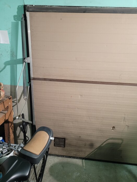 Garageport med cykel inuti förråd. Porten har skumisolering och förbättrad tätning efterfrågas för energibesparing. Prototyp-planer för byte nämns.