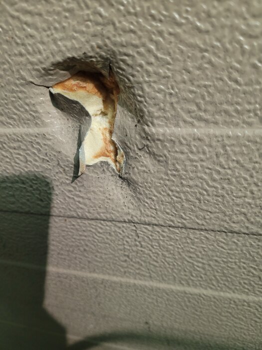 Närbild av en garageport med en skadad yta där skumisolering är synlig inuti genom ett sprucket hål.