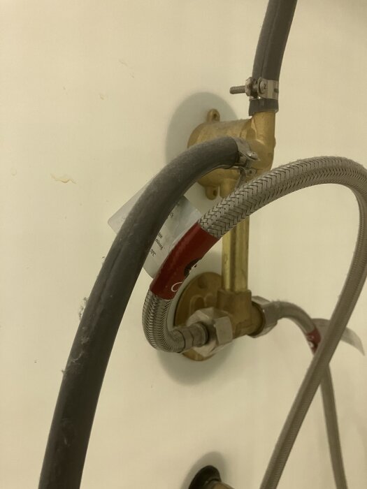 Duschens rör och slangar, med focus på kopplingar och en engreppsblandare. Syntetisk och metallisk slang mot en vit vägg.