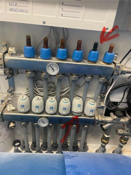 Bild på golvvärmefördelare med blå styrdon, ett markerat med röd pil, och vita termostatventiler i ett tekniskt skåp.