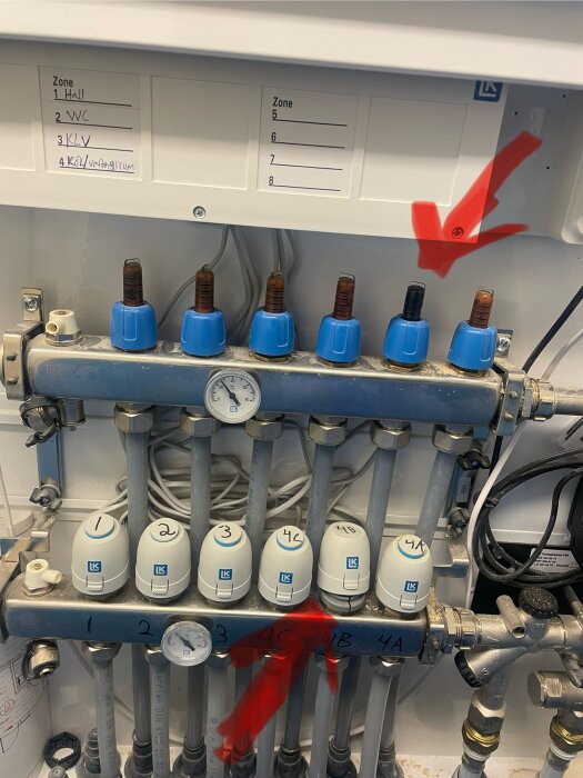 Golvvärmesystemets styrdon med flera anslutna rör och en lös enhet markerad med röda pilar.