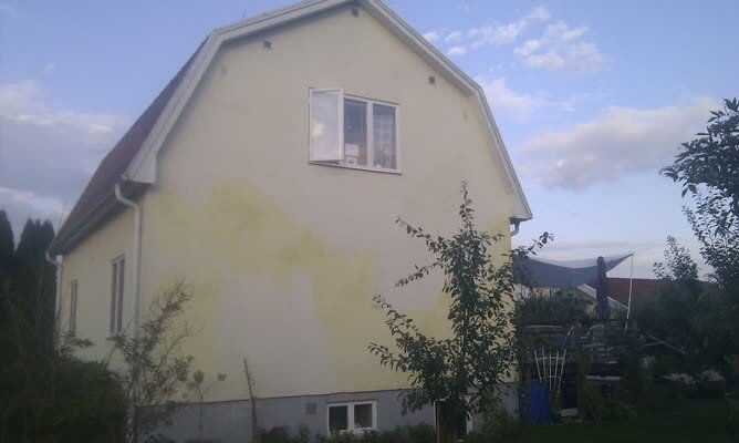 Gul villa med mansardtak och uppvuxen trädgård i förgrunden. Huset från 1939 har två fönster på gaveln och skadefläckar på fasaden.