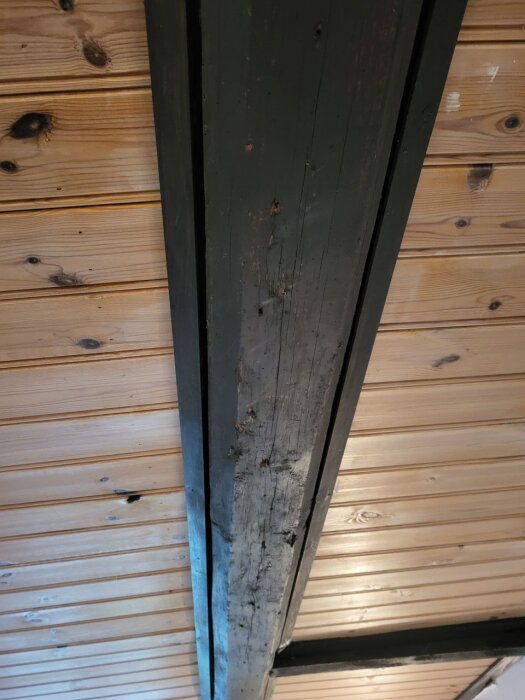 Mörk målad bjälke i trätak med ljusare träpanel i ett gammalt hus från tidigt 1900-tal.