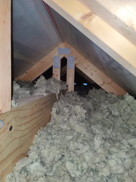 Isolerat utrymme i en vind med träkonstruktion och isoleringsmaterial; möjligt svartmögel synligt vid takstolsbalk.