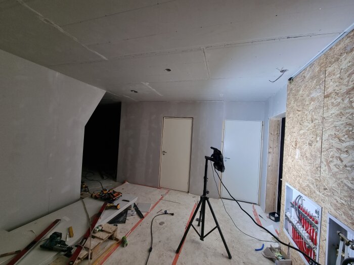 Rum under renovering med två dörrar, verktyg på golvet, och rörsystem synligt.