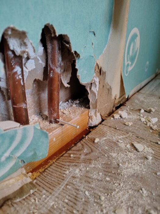 Skadat rör bakom en trasig vägg med vattenläckage och byggdamm på golvet i ett sovrum under renovering.