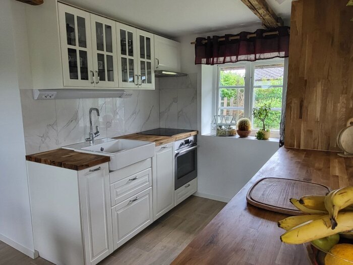 Kompakt kök med vita skåp, marmorerat kakel, fläkt ovanför spishällen, och träbänkskiva. Fönster med planterade växter.