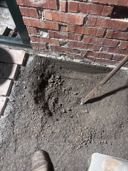 Hål i marken framför en tegelvägg, omgiven av grus och sten, med en sko och ett järnspett synliga i bild.