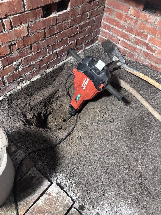 Bilmaskin Hilti och grävt hål i växthusets jordgolv, omgärdat av röd tegelvägg.