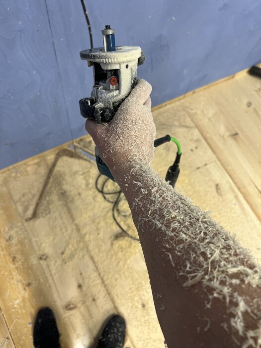 En person håller en handöverfräs, armen är täckt av träspån, på ett trägolv med spån.