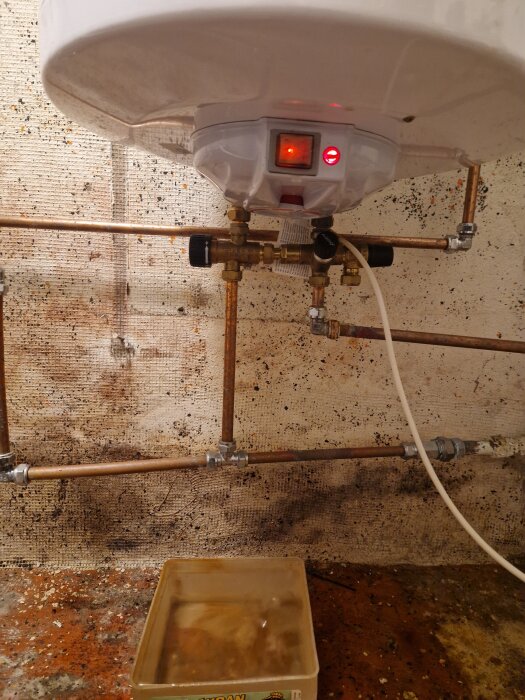 Värmesystem med rör och vattenbehållare på golvet. Enhet med röd indikator i mitten, dropp och rostfläckar syns på väggen bakom.