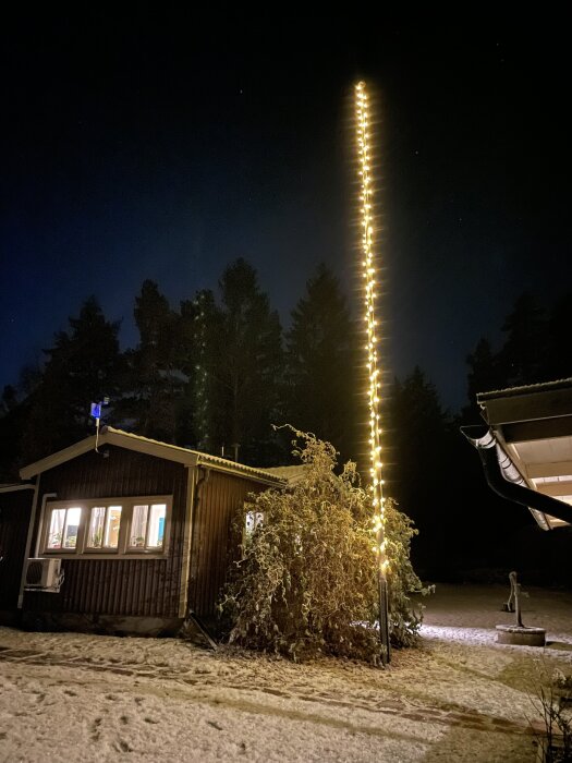 En flaggstång lindad med gulorange ljusslinga lyser upp natthimlen framför ett mörkt trähus omgiven av snö och träd i bakgrunden.