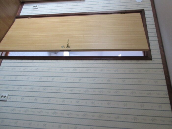 Solid dörr med träram och träfaner, monterad horisontellt i en vägg med randigt blommönstrat tapet. 80-tals design, okänt trädslag.